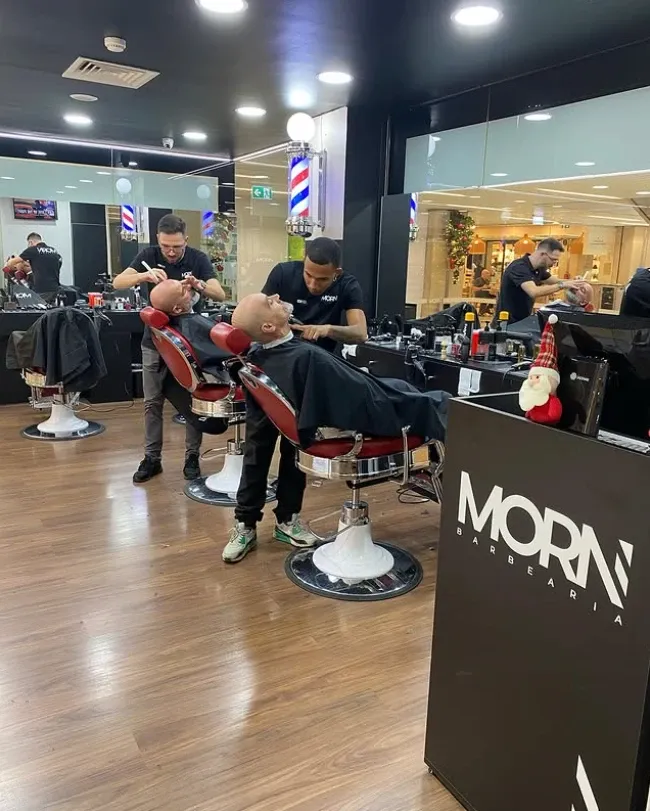 Interior da MORN Barbearia: dois barbeiros a fazer a barba a clientes nas cadeiras vermelhas, com o balcão MORN em primeiro plano