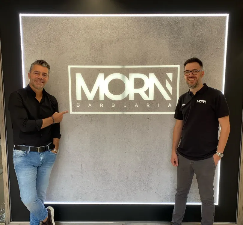 Duas pessoas junto ao painel iluminado com o logótipo da MORN Barbearia